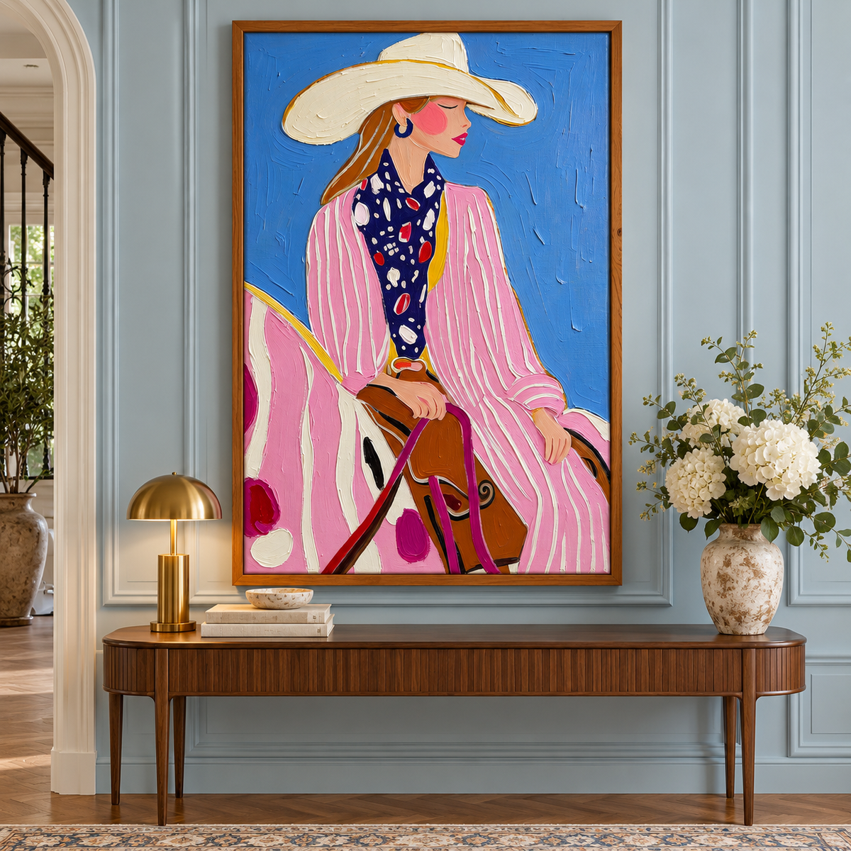 A cowgirl in a pink-and-white striped outfit, cream cowboy hat and dark patterned scarf seated on a pink-and-white horse against a bright blue background, shown in room setting 2.